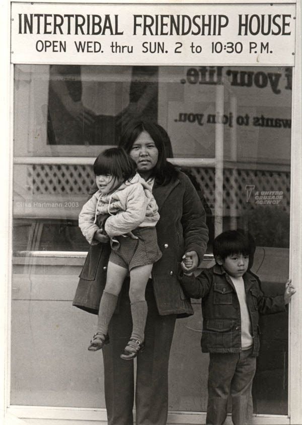 Black and white photograph featuring a Native American woman standing outdoors, wearing a thick coat, holding an agitated toddler in one arm and the hand of a young boy standing next to her. The text 'Intertribal Friendship House OPEN WED. thru SUN. 2 to 10:30 P.M.' is printed on the building behind them.